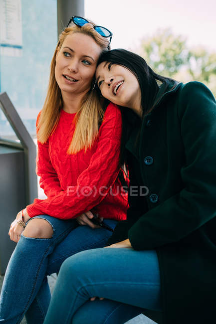 Jeune femme asiatique souriante et penchée sur l'épaule d'un ami caucasien assis sur un banc et attendant le bus ensemble — Photo de stock