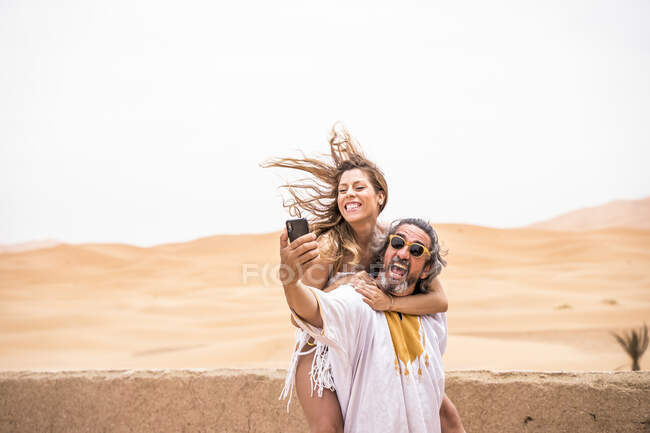 Middle-aged man with woman piggyback taking selfie expressively on terrace against sandy desert, Morocco — Stock Photo