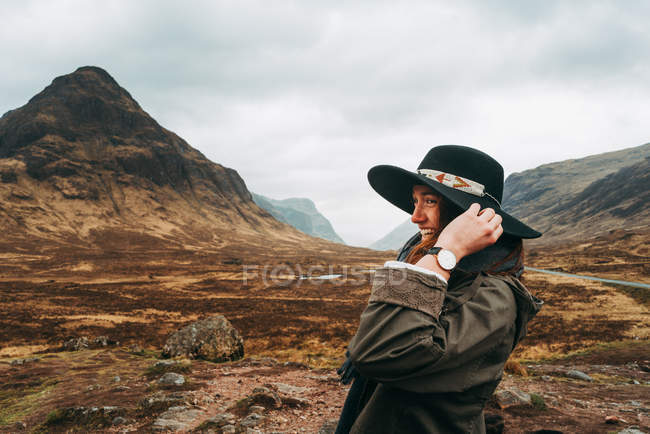 Beautiful ginger woman wearing and holding hat standing against picturesque mountains of Scotland — Stock Photo