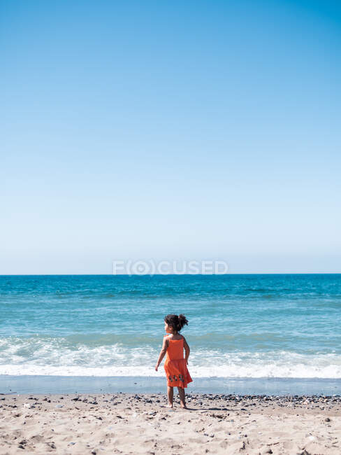 Vista posteriore del bambino femmina in abito arancione in piedi sul mare sabbioso e guardando mare e cielo — Foto stock
