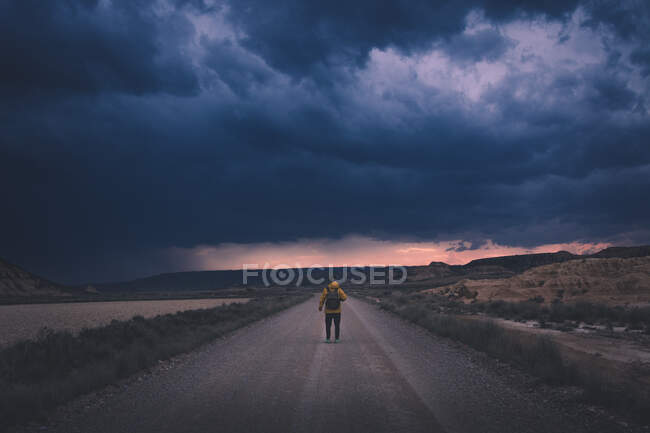 Vista trasera de un joven con chaqueta amarilla y mochila de pie en un camino vacío que se extiende alto hasta la noche sombría nubes impresionantes en el semi-desierto Bardenas Reales Navarra España - foto de stock
