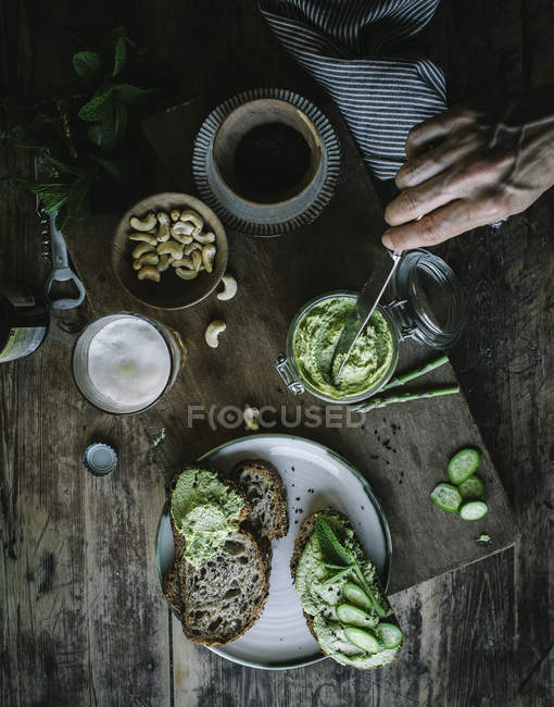 Сверху рука неузнаваемого человека, делающего тосты с зеленым кешью и ломтиками огурца на деревянной доске — стоковое фото