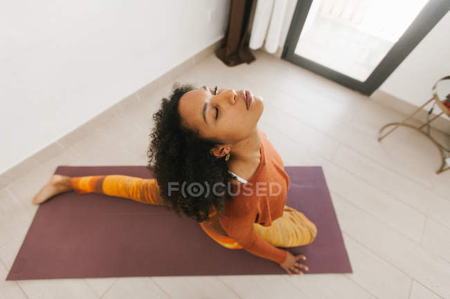 Rosto da jovem mulher afro-americana sentada em pose de ioga com olhos fechados no tapete na sala de luz — Fotografia de Stock