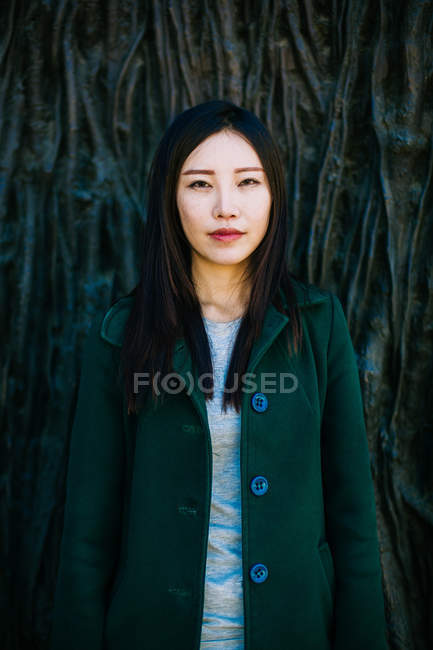 Pensive mulher asiática na roupa da moda sorrindo e olhando para longe, enquanto se inclina na parede com alívio de raízes de árvores — Fotografia de Stock