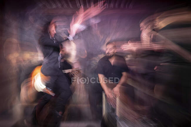 Homme en costume noir dansant le flamenco près des musiciens hispaniques masculins pendant la représentation contre la peinture sur scène sombre — Photo de stock