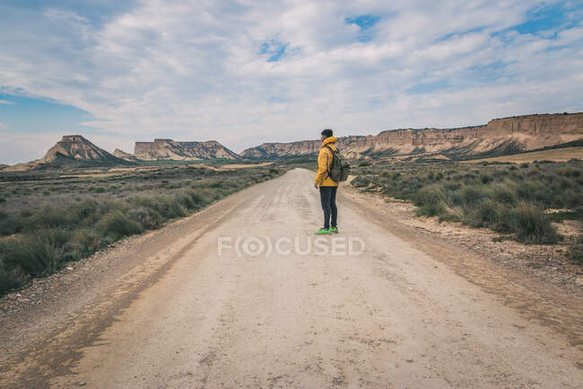 Vista lateral do jovem de jaqueta amarela e mochila em pé na estrada vazia esticando-se alto entre colinas pedregosas no semi-deserto Bardenas Reales Navarra Espanha — Fotografia de Stock