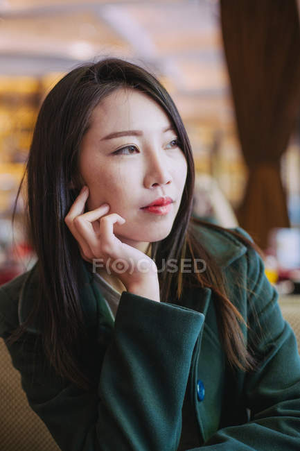 Азиатка в стильном зеленом пальто смотрит вдаль, сидя за столиком кафе — стоковое фото