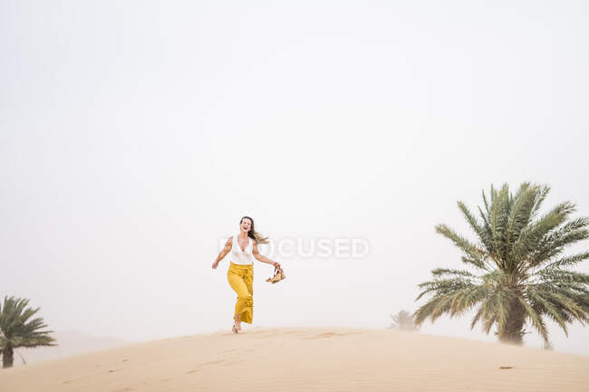 Cheerful stylish blonde woman holding shoes while walking in desert of Morocco — Stock Photo