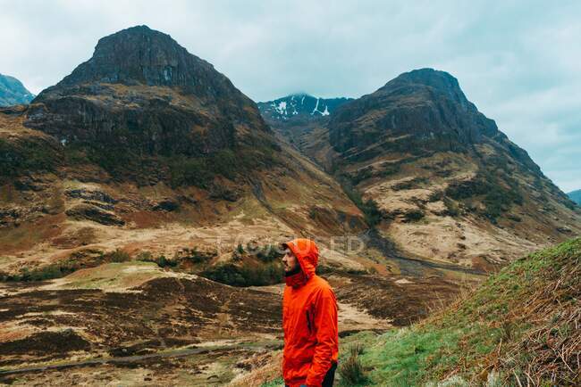 Vista lateral do jovem vestindo um casaco vermelho com capuz enquanto está de pé contra montanhas pitorescas da Escócia — Fotografia de Stock