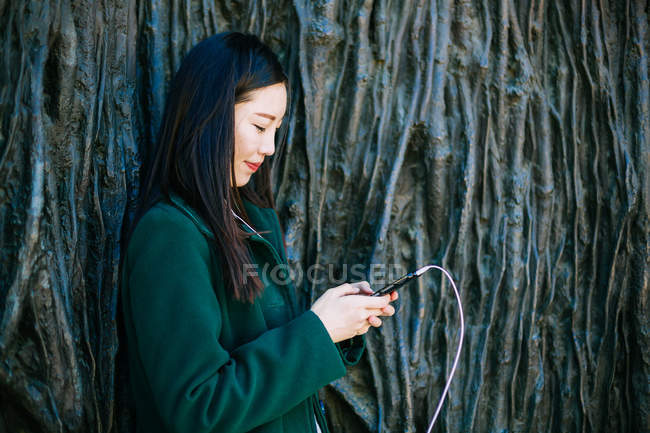 Красивая азиатка в стильном наряде слушает музыку и использует смартфон, опираясь на грубую стену с рельефом деревьев — стоковое фото
