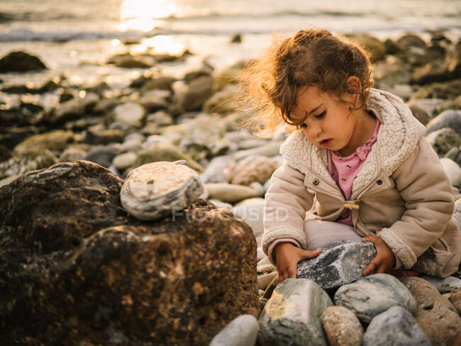 Nettes interessiertes Kleinkind, das mit Stein spielt, während es am Meer sitzt — Stockfoto