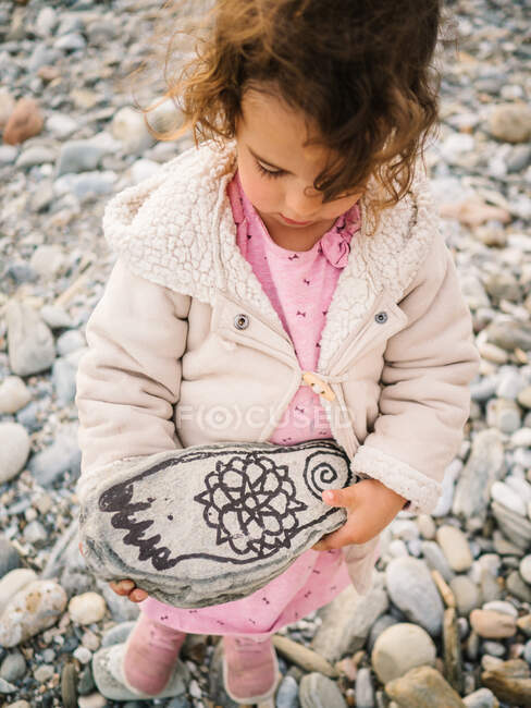 D'en haut mignon intéressé tout-petit fille jouer avec pierre peinte sur bord de mer — Photo de stock