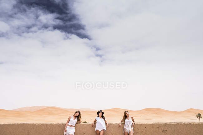 Ряд женщин в белой пляжной одежде, позирующих у каменного забора на террасе против бесконечной пустыни, Марокко — стоковое фото
