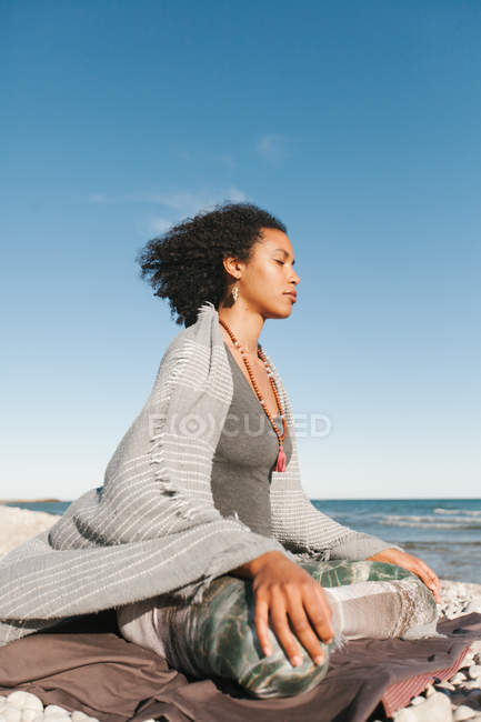 Mujer joven afroamericana meditando en la postura de loto yoga en la playa de arena en un día brillante - foto de stock