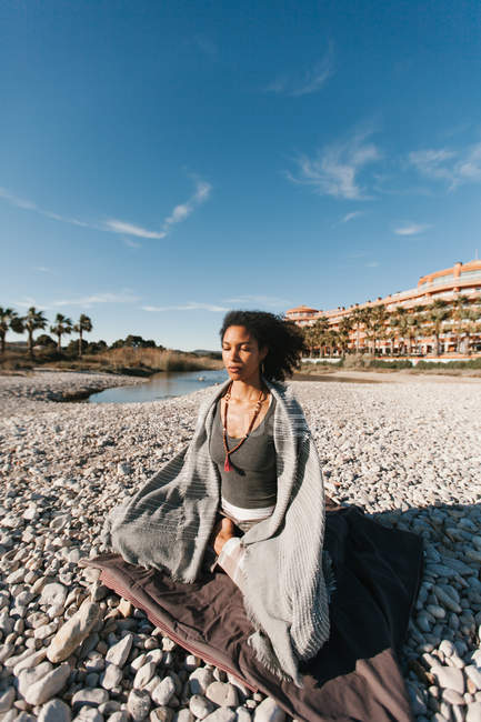Привлекательная молодая женщина медитирует и молится в ошейнике джапамалы в позе йоги лотоса на песчаном пляже в яркий день — стоковое фото