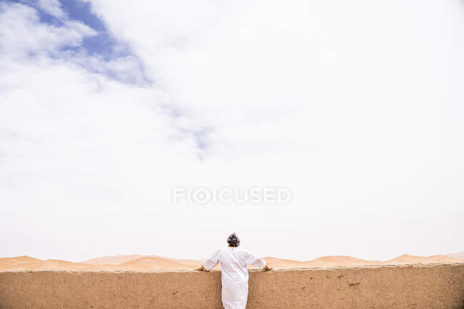 Rückansicht eines anonymen erwachsenen Mannes in langen weißen Kleidern, der sich an eine Wand lehnt und wegblickt gegen die endlose Sandwüste, Marokko — Stockfoto