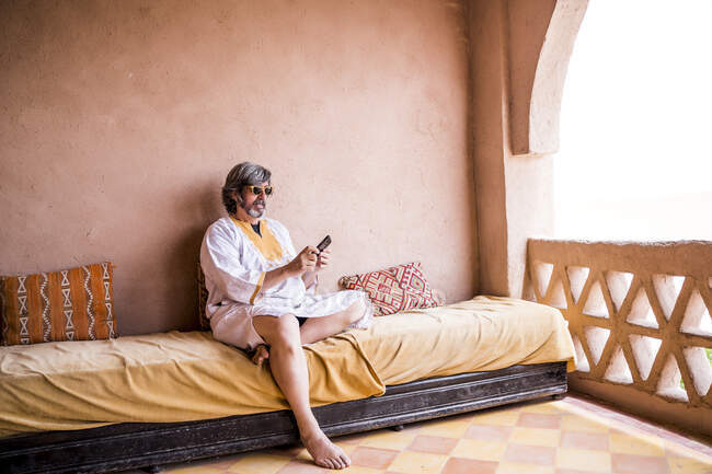 Hombre adulto en ropa larga sentado en el sofá en la terraza con valla de piedra en estilo oriental y el uso de teléfono, Marruecos - foto de stock