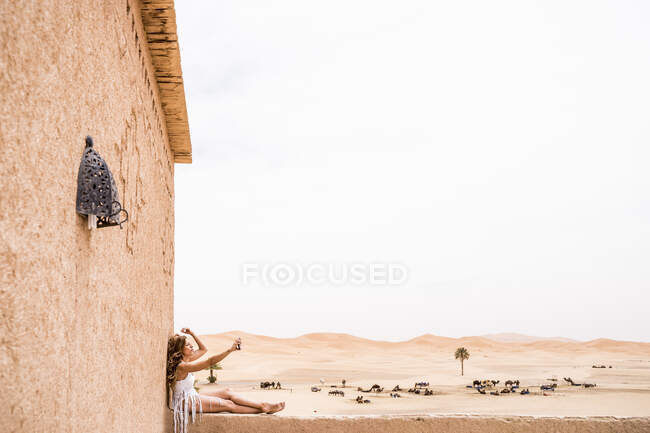 Vista lateral de la hermosa joven en la parte superior blanca sentada en la valla de piedra en el viento tomando una selfie contra el desierto de arena sin fin, Marruecos - foto de stock