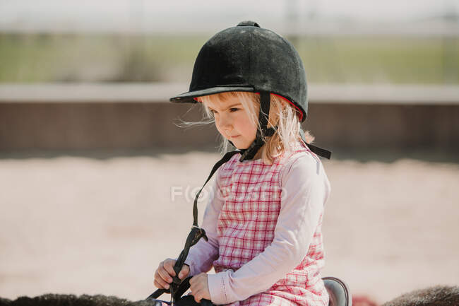 Kleines Mädchen in Kleid und Jockey-Heu sitzt auf dem Pferd und lernt auf der Rennbahn zu reiten — Stockfoto