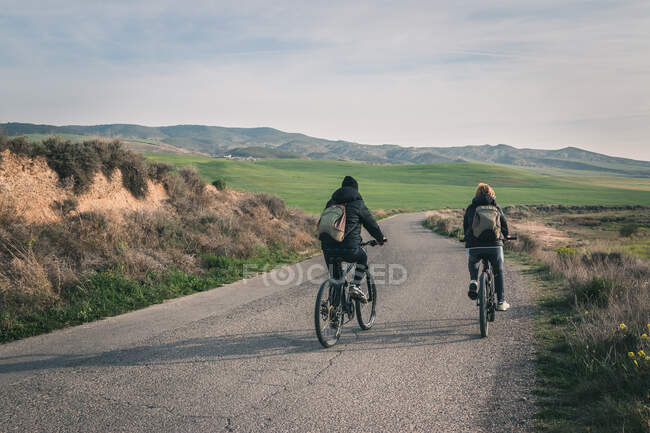 Back view of young men in dark clothes and backpack riding bicycles on empty road winding between stony hills in semi-desert Bardenas Reales Navarra Spain - foto de stock