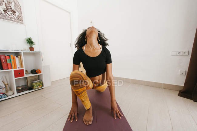Femme afro-américaine assise dans une pose de yoga sur un tapis dans une pièce lumineuse — Photo de stock