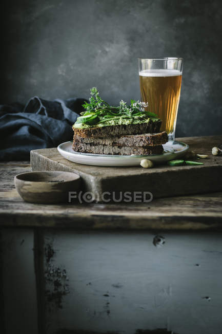 Toasts with green cashew pate, herbs and slices of cucumber with glass of beer on wooden board — Stock Photo