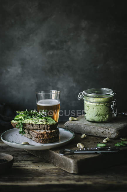 Тосты с зеленым кешью паштет, травы и ломтики огурца с банкой и стаканом пива на деревянной доске — стоковое фото