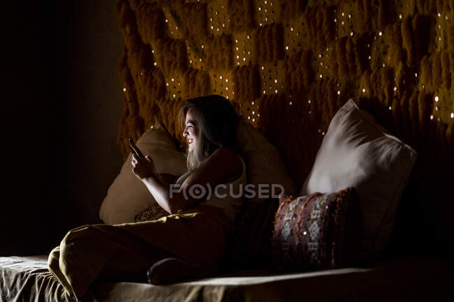 Side view of young lady smiling and using smartphone while sitting on soft bed in dark room in Morocco — Stock Photo