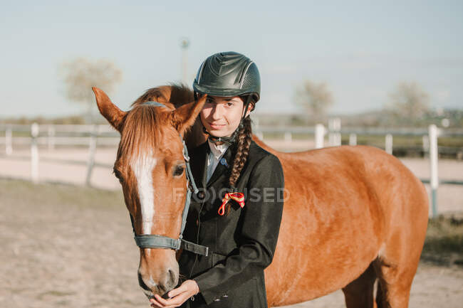 Vista lateral de joven adolescente mujer en casco de jinete y chaqueta acariciando caballo de pie juntos al aire libre y mirando a la cámara - foto de stock