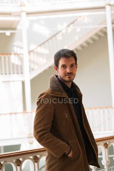 Erwachsener Mann in brauner Jacke mit Händen in Taschen steht in heller Halle mit Treppe und blickt in die Kamera — Stockfoto