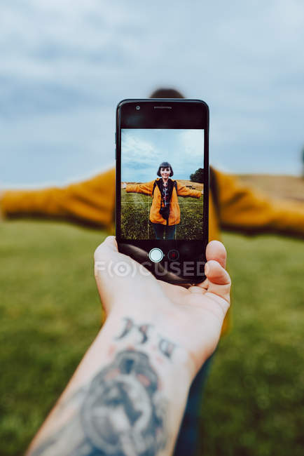 Ritaglia l'immagine dell'uomo tatuato usando lo smartphone per fotografare la giovane donna con le braccia tese in campo verde — Foto stock