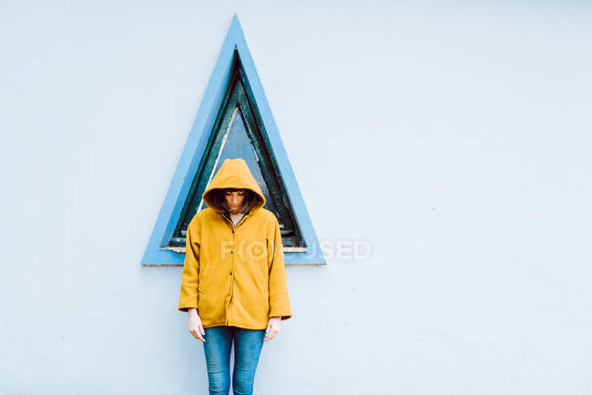 Молода жінка в жовтій теплій шубці посміхається і дивиться вниз, стоячи біля вікна трикутника і сірої стіни будівлі — стокове фото