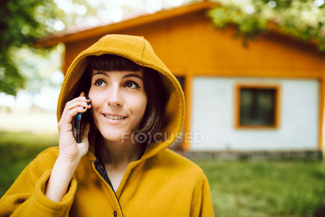 Junge Frau in lässigem Outfit lächelt und spricht per Smartphone, während sie auf gefliestem Pfad vor dem schönen Ferienhaus steht — Stockfoto