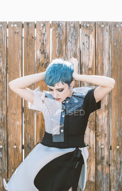 Young woman with short blue hair wearing futuristic dress and looking down while standing near shabby lumber fence on city street — Stock Photo