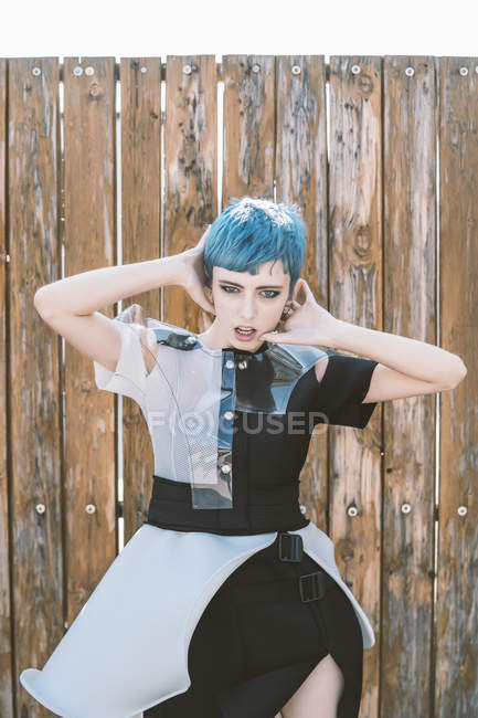 Young woman with short blue hair wearing futuristic dress and looking at camera while standing near shabby lumber fence on city street — Stock Photo