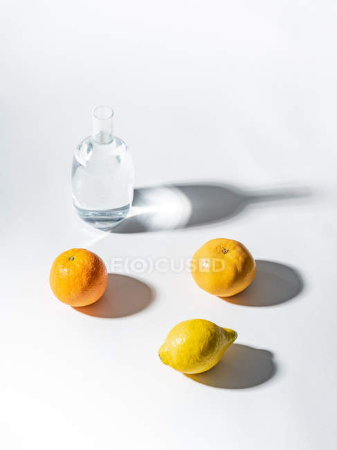 Стиглі апельсини та лимон біля прозорої банки з водою на білому тлі — стокове фото
