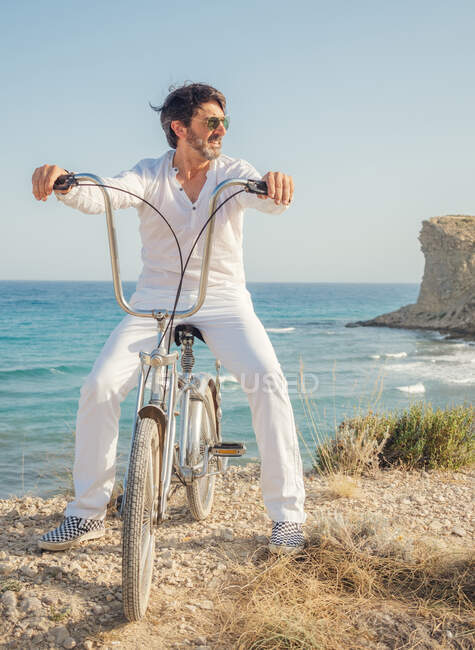 Mature bearded sporty man using sunglasses while biking on seaside with dry grass on background of amazing turquoise seascape in bright day — Stock Photo