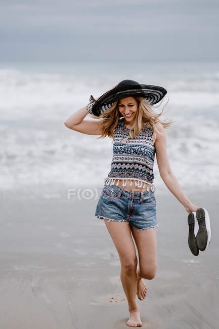 Привлекательная женщина в черной шляпе, держащая туфли во время прогулки по пляжу — стоковое фото