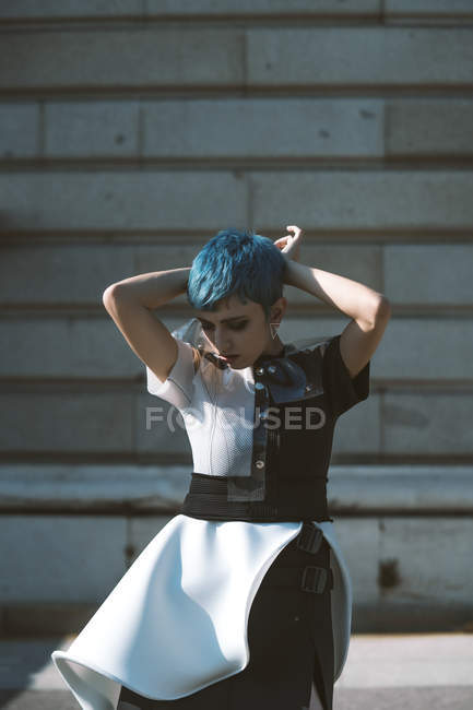 Молодая женщина в футуристическом платье трогает голову и короткие голубые волосы, стоя возле здания в солнечный день на городской улице — стоковое фото