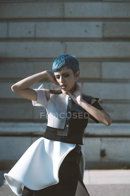 Young woman in futuristic dress touching face and short blue hair while standing near building on sunny day on city street — Stock Photo