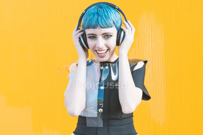 Fröhliche junge Frau im trendigen alternativen Kleid lächelt und hört Musik mit Kopfhörern gegen die gelbe Wand — Stockfoto