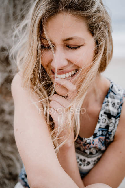 Mujer rubia en la parte superior mordiendo dedo índice y sonriendo con los ojos cerrados en el fondo de la naturaleza - foto de stock