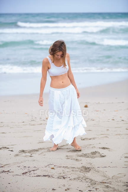 Atractiva hembra en traje blanco bailando en la arena cerca del mar ondulante - foto de stock