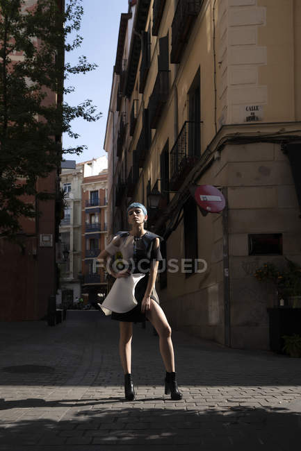 Young woman in futuristic dress standing with hands on waist on street against old building in sunlight — Stock Photo