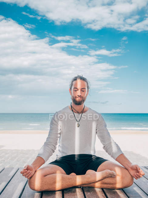 Взрослый бородатый мужчина медитирует, сидя в позе лотоса на деревянном пирсе у берега моря с скрещенными ногами и закрытыми глазами — стоковое фото