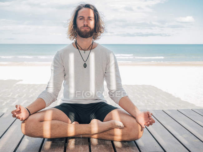 Homme barbu adulte méditant assis dans la pose de lotus sur une jetée en bois au bord de la mer avec les jambes croisées et regardant la caméra — Photo de stock