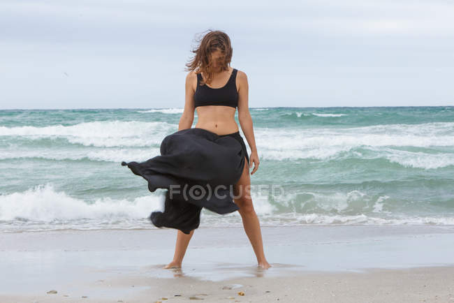 Привлекательная женщина в черном наряде танцует на песке возле моря — стоковое фото