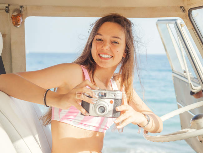 Femme en maillot de bain avec caméra souriant et prendre des photos en blanc siège avant de la voiture au bord de la mer dans la journée ensoleillée en regardant la caméra — Photo de stock