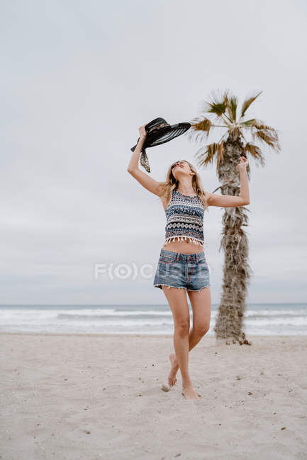 Attractive woman wearing top and shorts dancing on sandy seashore with black hat in raised hand — Stock Photo