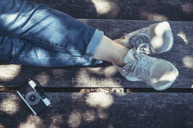 Von oben gekreuzte Beine in bequemen Turnschuhen und Fotokamera auf Holzbank bei strahlendem Sonnenschein — Stockfoto